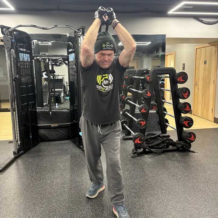 Man in a Body By Michelle beanie and T-shirt lifting hand weights overhead
