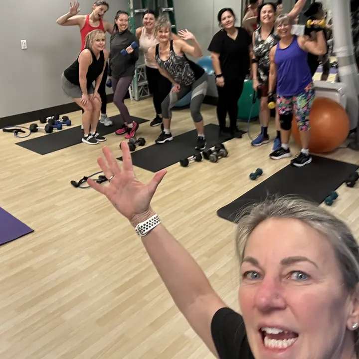 Group class waving to the camera, with mats and hand weights on the floor