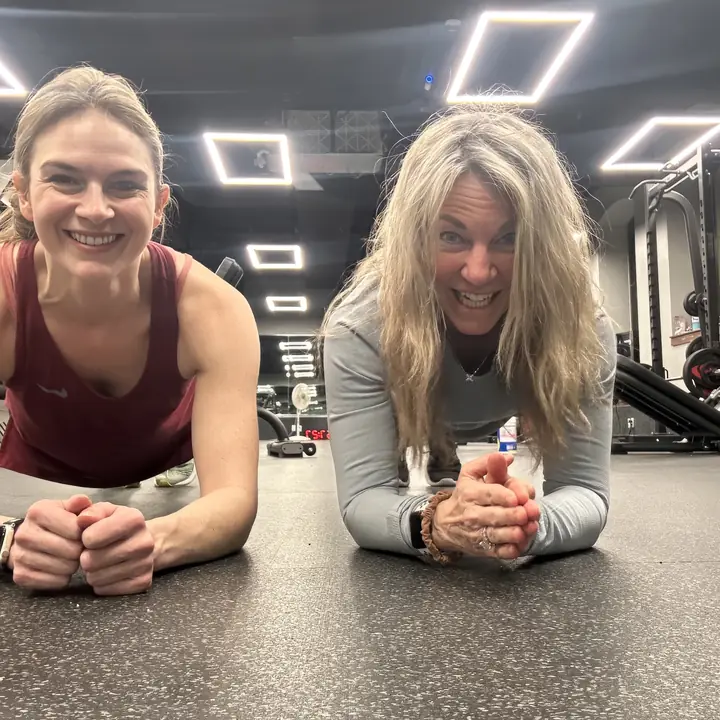 Two women holding a plank on the gym floor, smiling at the camera