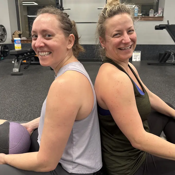 Two women sitting back to back on the gym floor, laughing